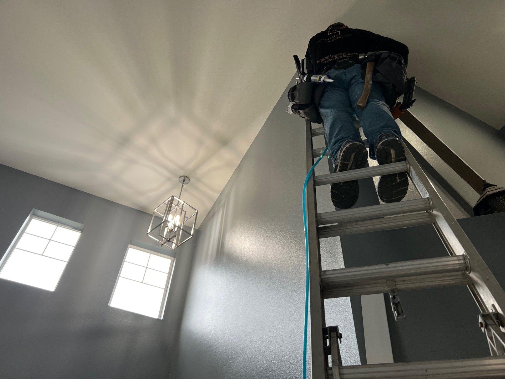 Person on ladder painting a gray wall. Light fixture, windows, and tools visible.