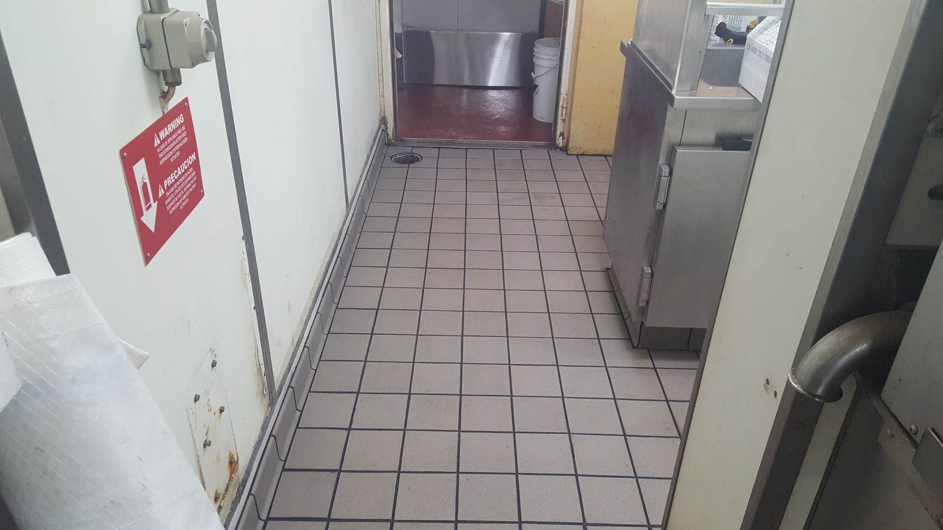 Narrow kitchen aisle with worn tile floor, stainless steel appliances, and a fire exit sign on the wall.