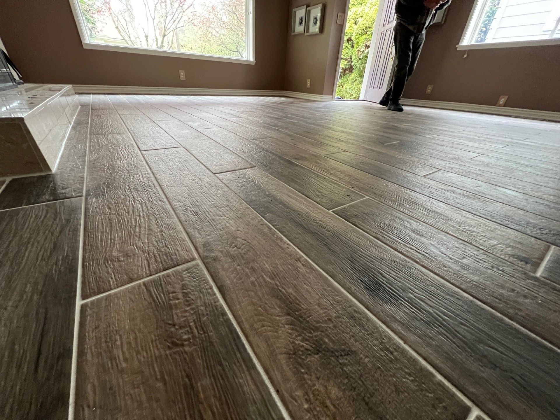 Close-up view of wood-look floor tiles being installed in a room with windows; worker standing.