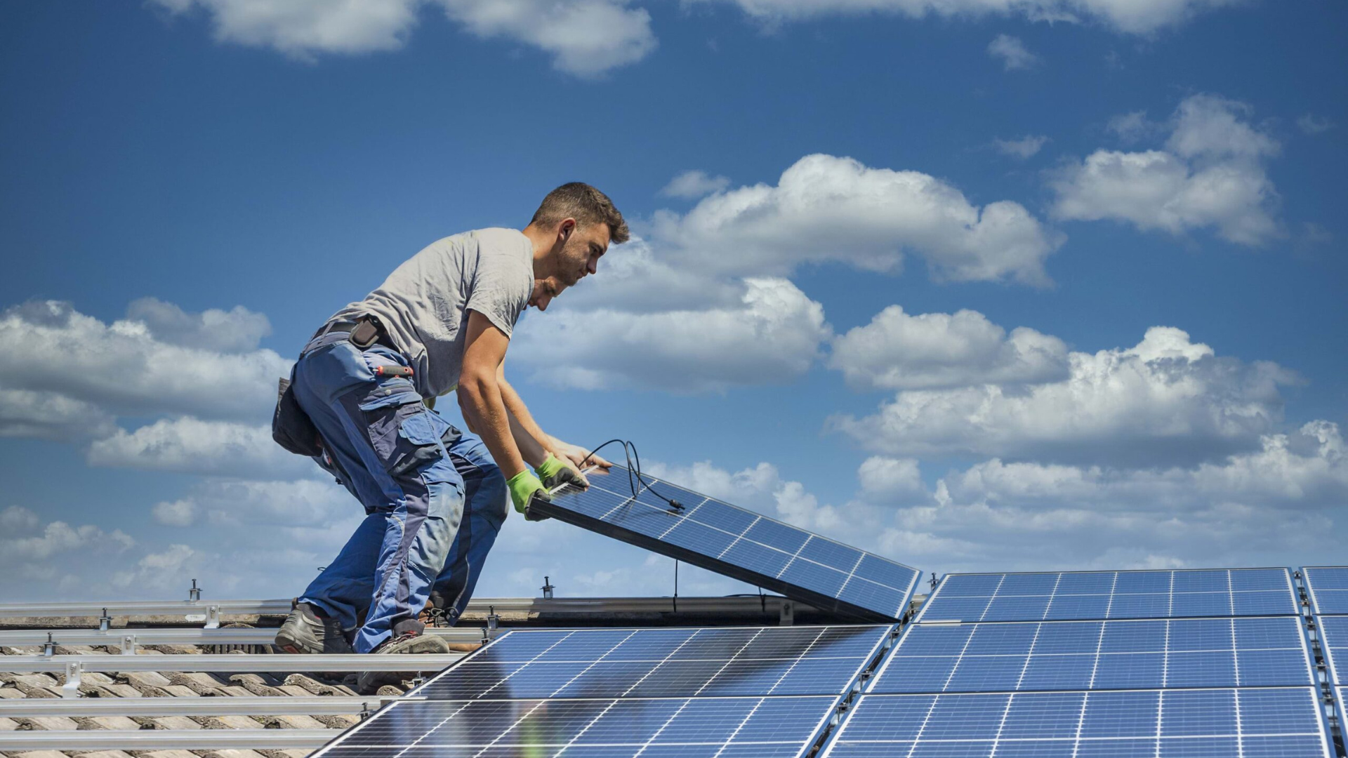Man installing solar panel on rooftop against a blue sky with clouds.