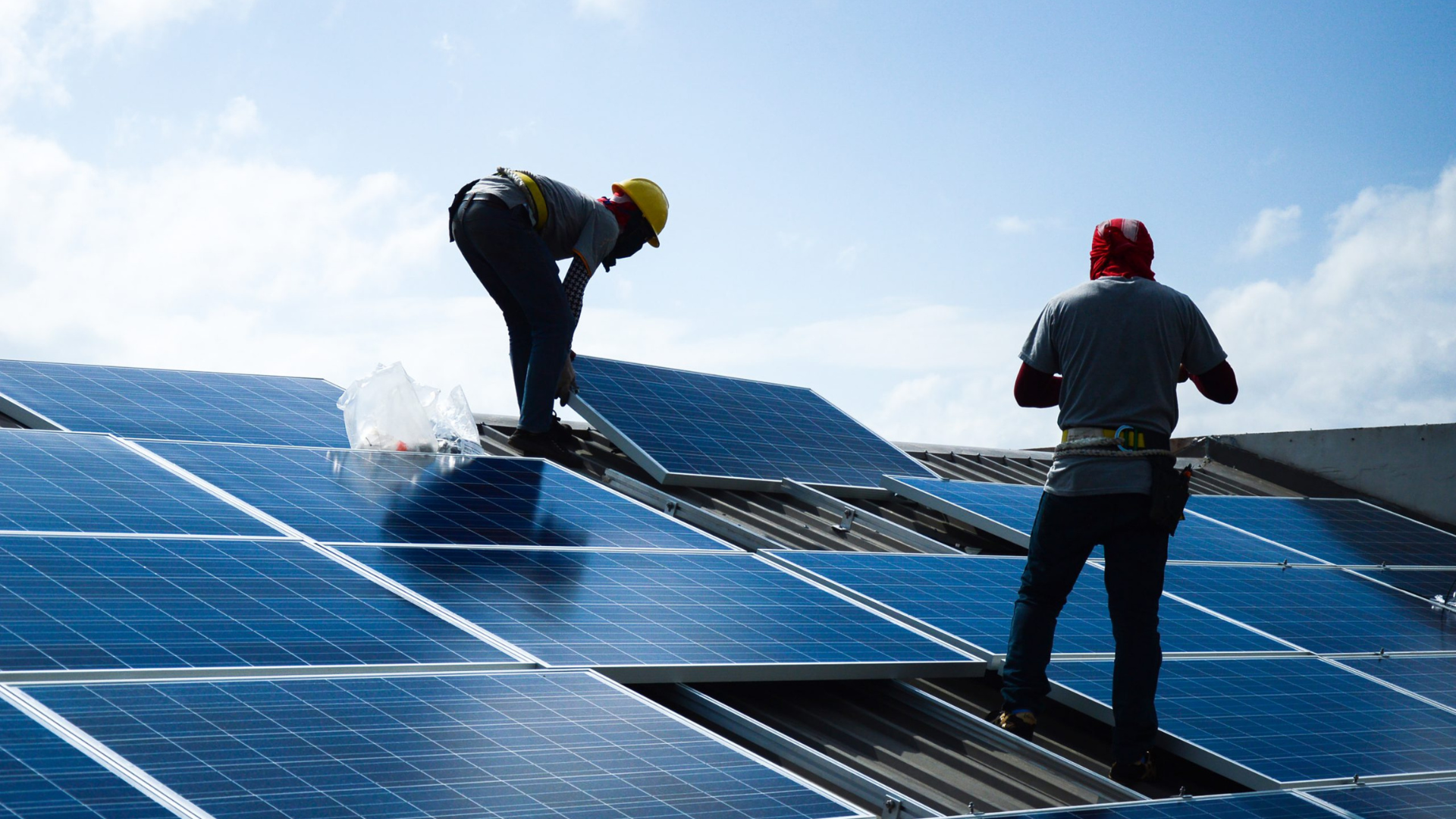 Two workers installing solar panels on a rooftop under a bright blue sky.