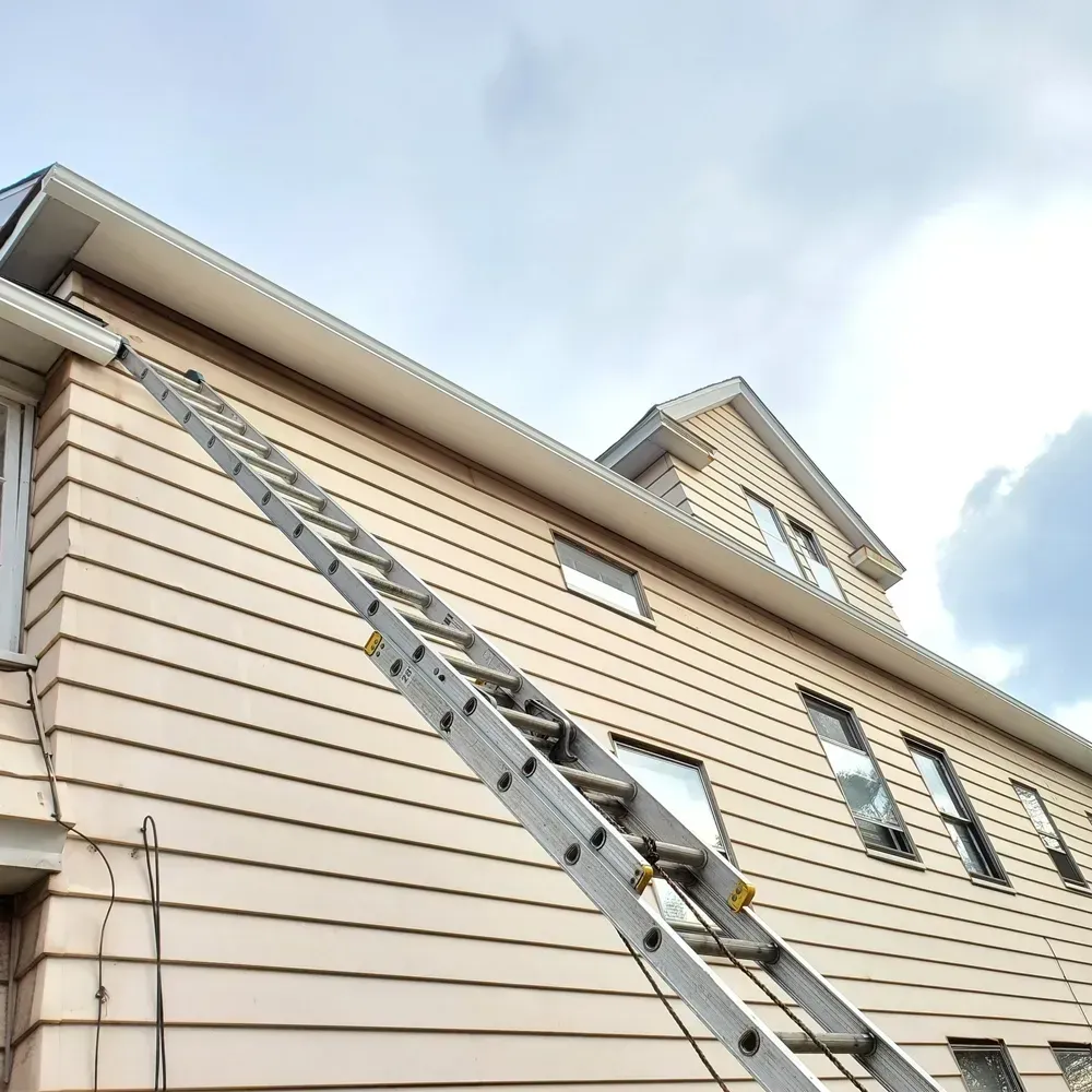 An aluminum extension ladder leans against the side of a light-colored, two-story house with beige siding.