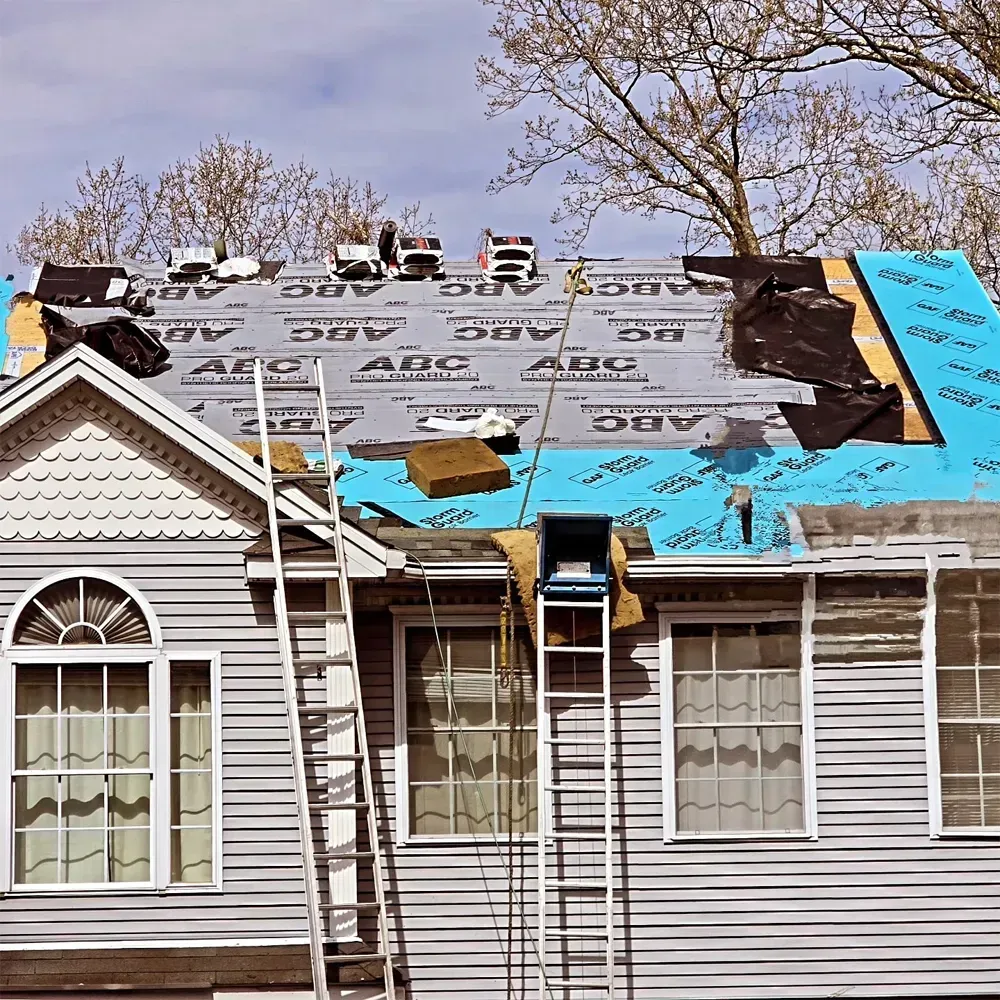 A roof under renovation with gray and blue underlayment, two ladders leaning against the house exterior.