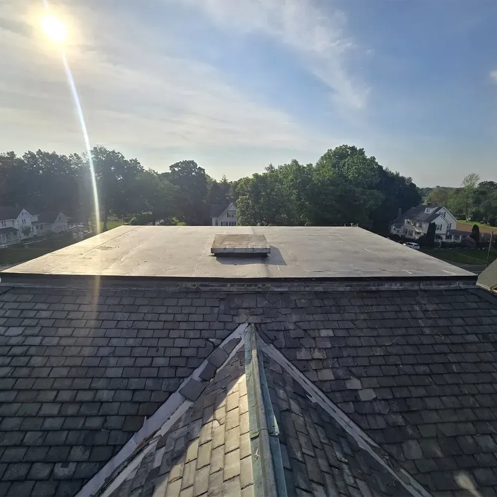 An elevated view of a flat roof section with a small skylight, rising above a lower pitched slate shingle roof.