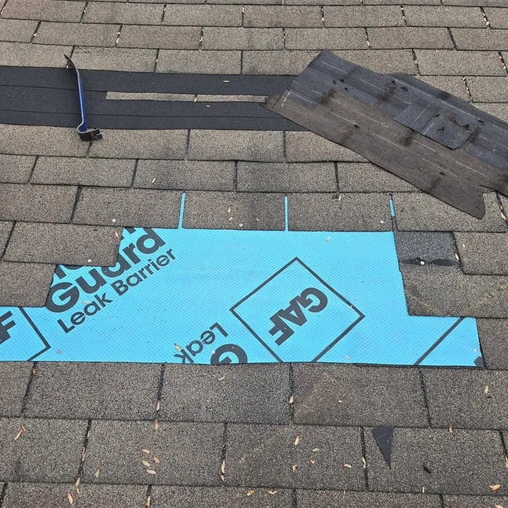 A roof under repair with a blue GAF leak barrier patch exposed, a pry bar, and removed shingles nearby.