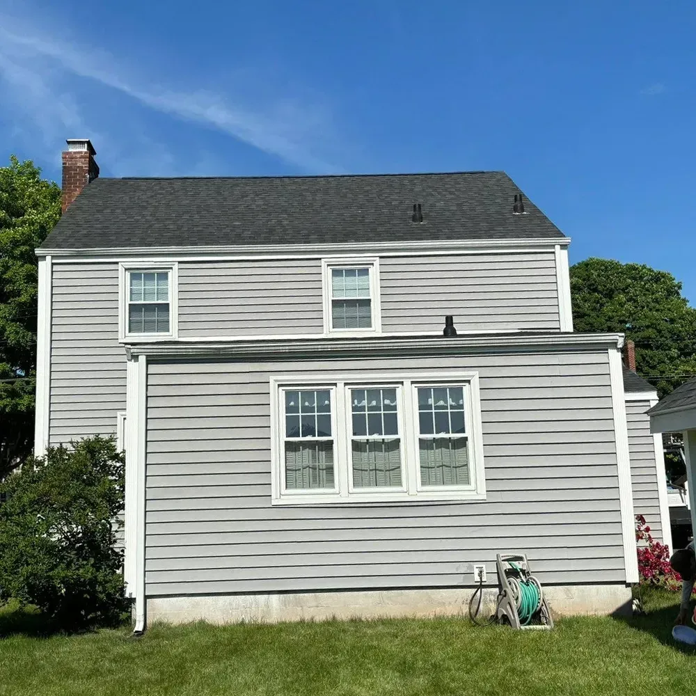 A light gray, two-story house with horizontal siding and a dark shingled roof stands against a bright blue sky.