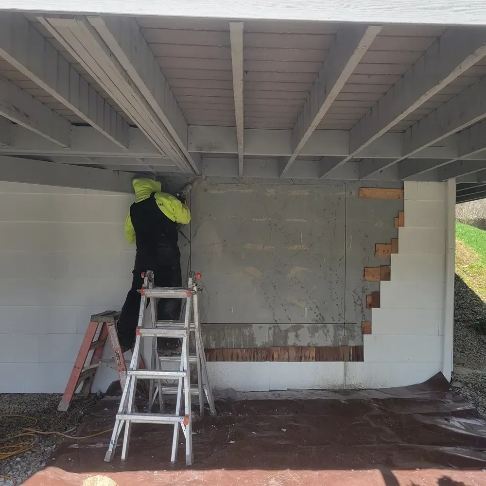 A worker in a yellow jacket stands on a ladder, installing panels on the gray wall under a wooden deck.