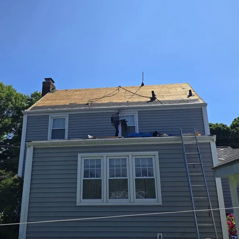A two-story house with blue siding under a clear blue sky, featuring a roof under repair with exposed wooden sheathing.