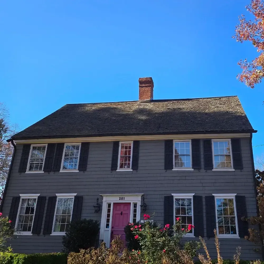 A two-story grey colonial house with black shutters, a vibrant pink front door, and a brick chimney under a blue sky.
