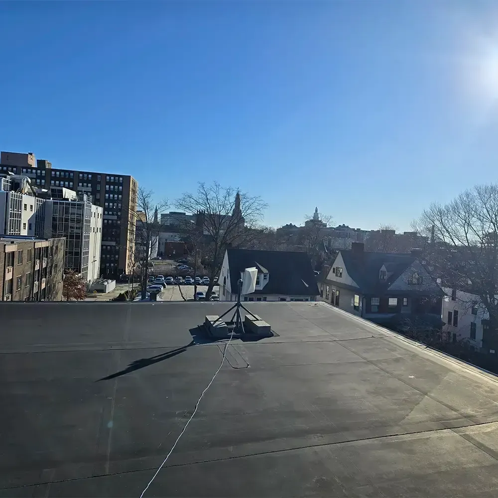 A roof view showing a wireless antenna unit on a weighted stand against a bright blue sky above city buildings.