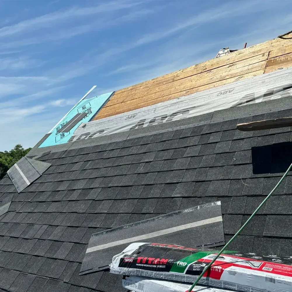 A partially shingled roof under a clear blue sky, showing exposed wood decking, roofing paper, and a pack of shingles.