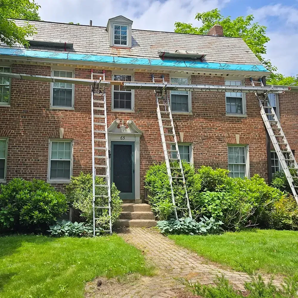 A two-story brick house under renovation, featuring ladders against the facade and blue weatherproofing on the roof.