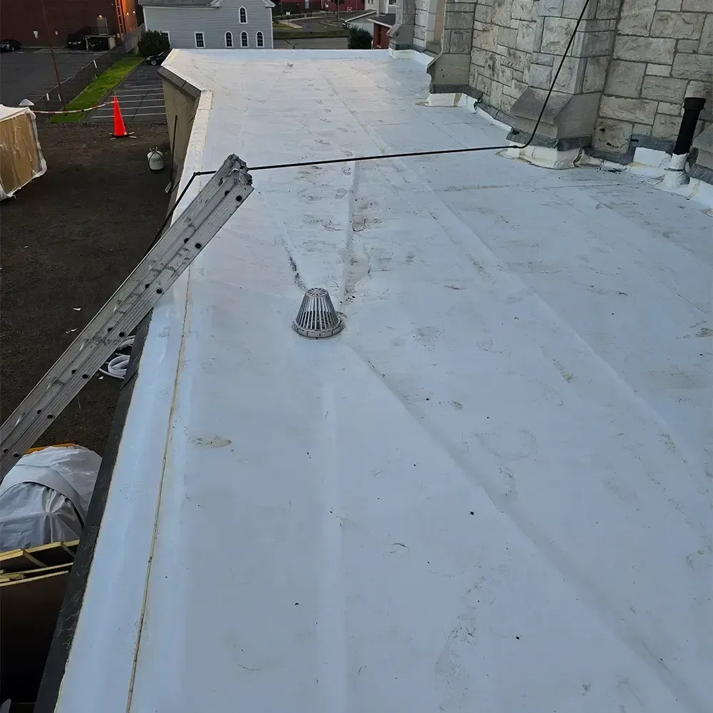 A white flat roof with a drainage dome, viewed from a ladder, with stone wall siding on the right and a parking lot below.