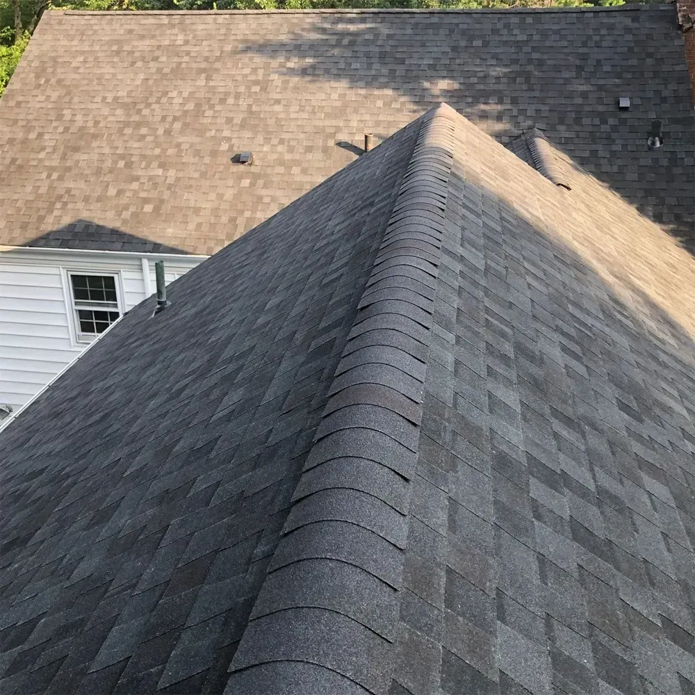 A high-angle view of a gray shingled roof with a prominent ridge cap, looking down towards a white-sided building.