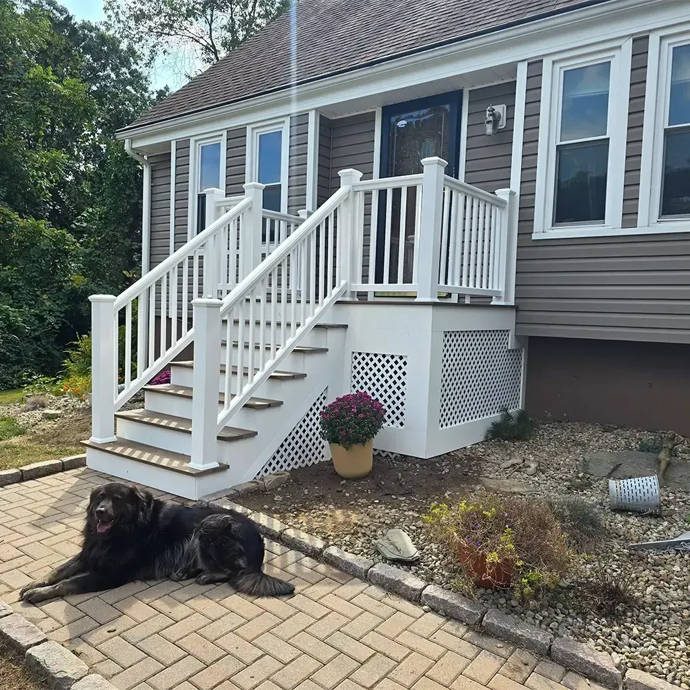 A large black dog lies on a herringbone brick patio in front of a house with white stairs and lattice skirting.