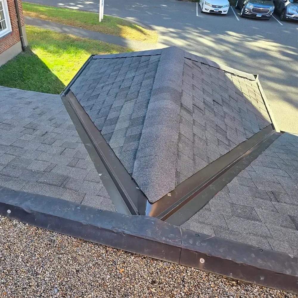 A high-angle view of a grey shingled roof extension with metal flashing, adjacent to a flat, gravel-covered roof.