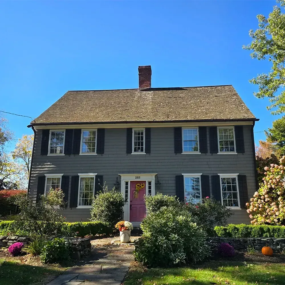 A two-story grey colonial house with a dark shingled roof, black shutters, and a bright pink front door under a clear sky.