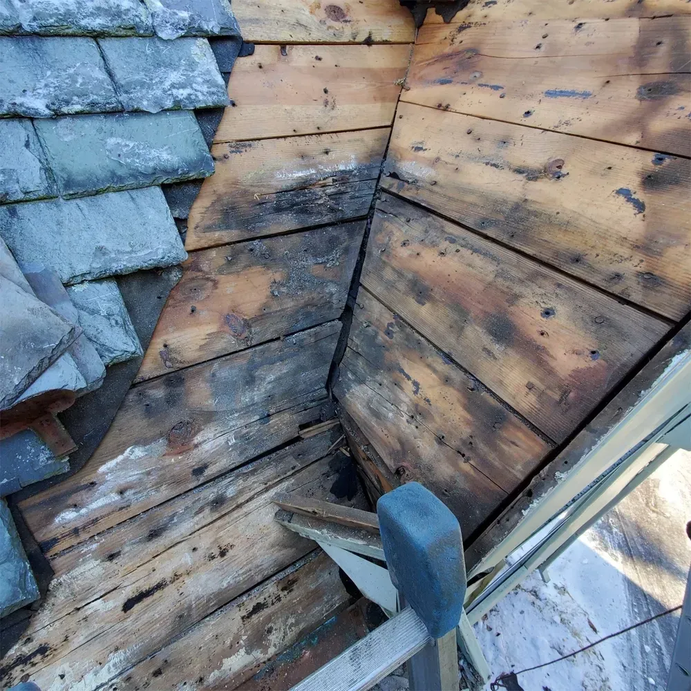 Exposed, weather-stained wood roof decking in a valley transition, adjacent to a section of old slate roofing tiles.