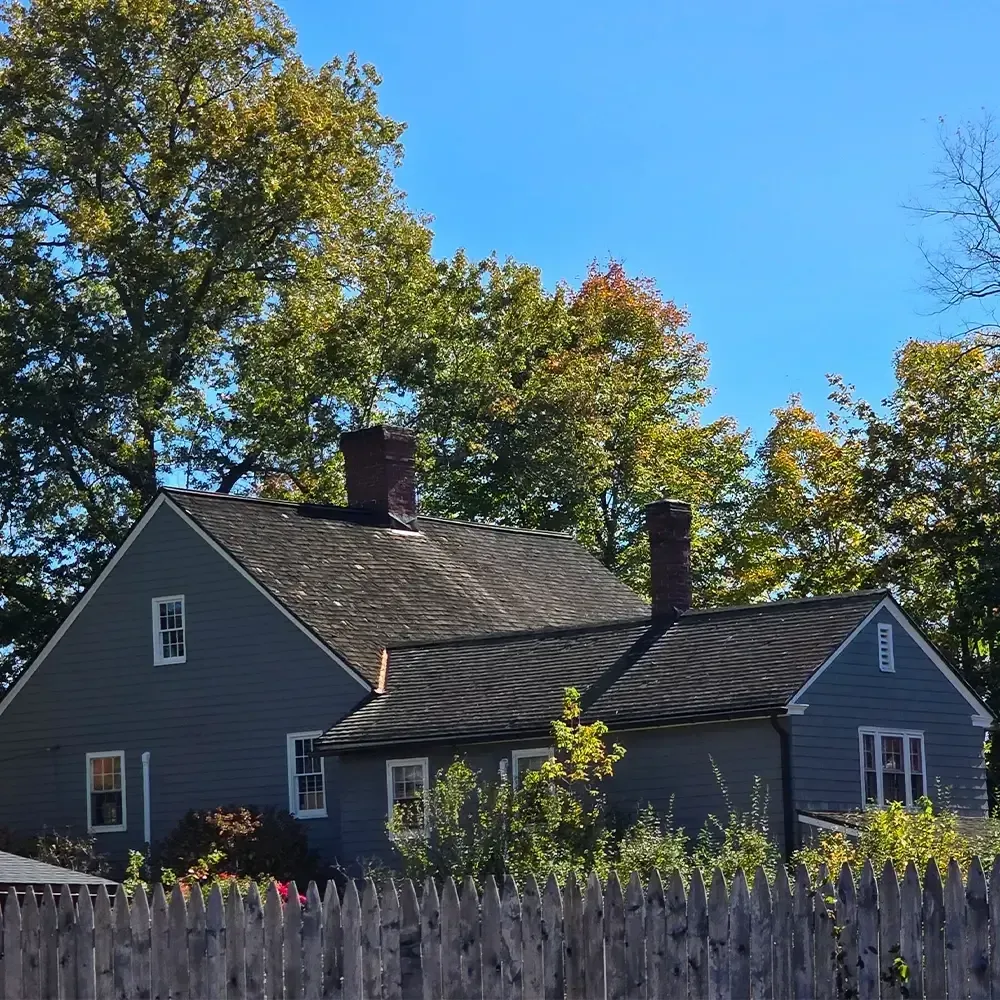 A gray, traditional-style house with two chimneys and a dark shingled roof, set against trees under a clear blue sky.