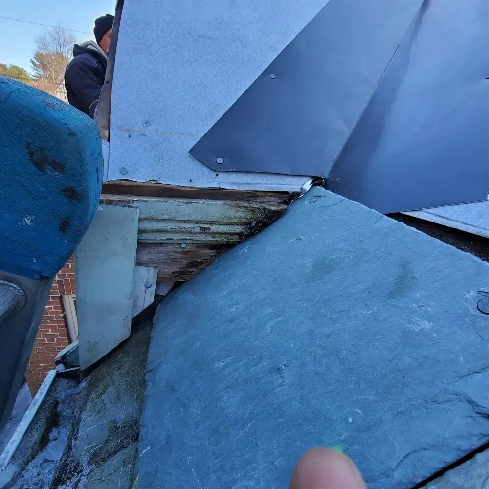 A close-up view of a roof repair site, showing exposed damaged wood trim beneath grey metal flashing and slate tiles.