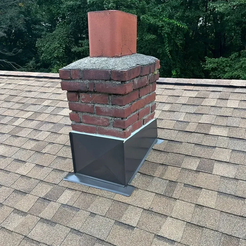 A brick chimney on a shingled roof with new metal flashing installed at the base.
