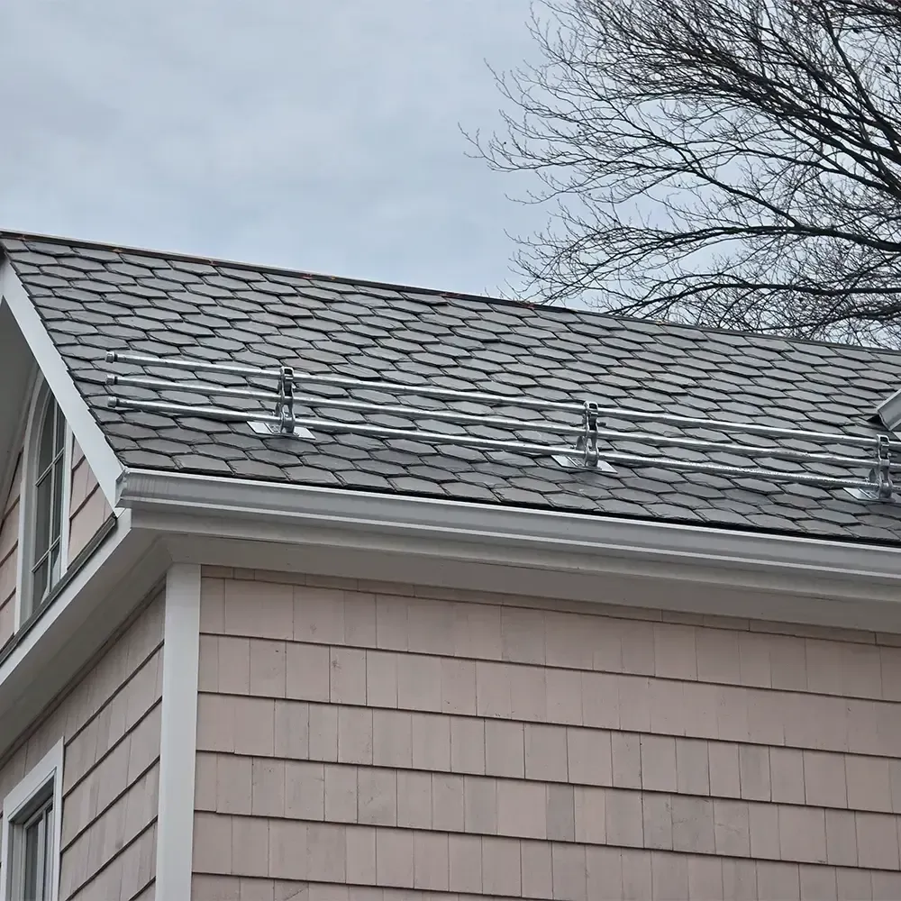 A metal snow guard rail installed on the dark gray shingled roof of a house with tan exterior siding.