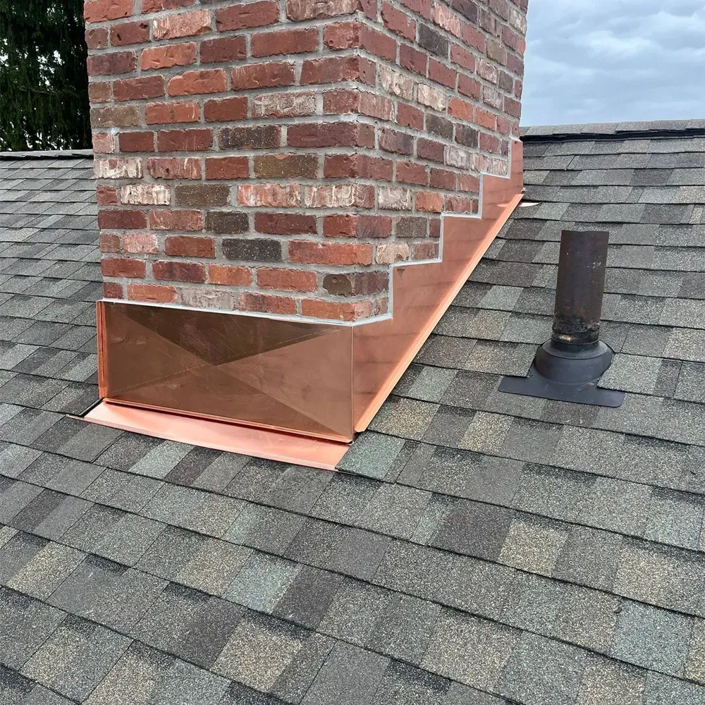 A brick chimney on a shingled roof features newly installed copper flashing around its base and an adjacent pipe vent.