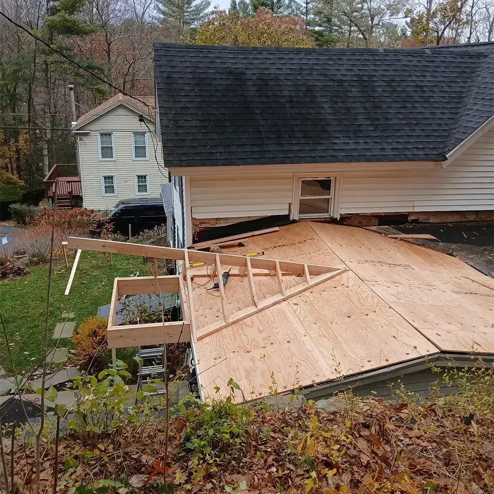 An outdoor view showing a wooden roof addition under construction attached to the side of a white house.