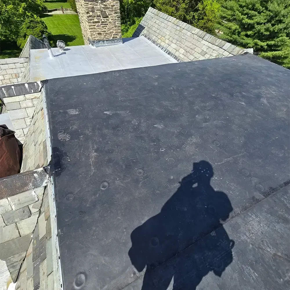 A high-angle view of a roof with a large black membrane section next to a light-colored flat roof and stone masonry.
