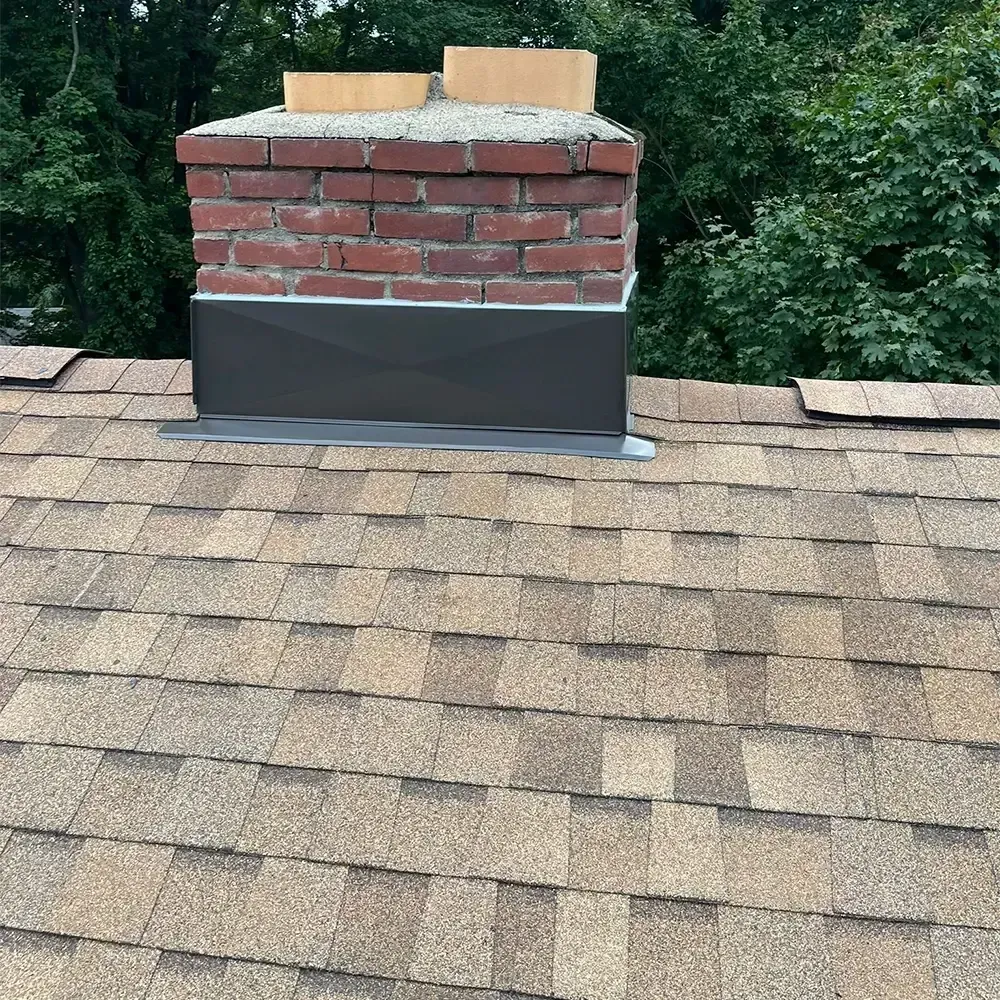 A brick chimney with a dark metal base flashing on a shingled residential roof, set against a background of green trees.