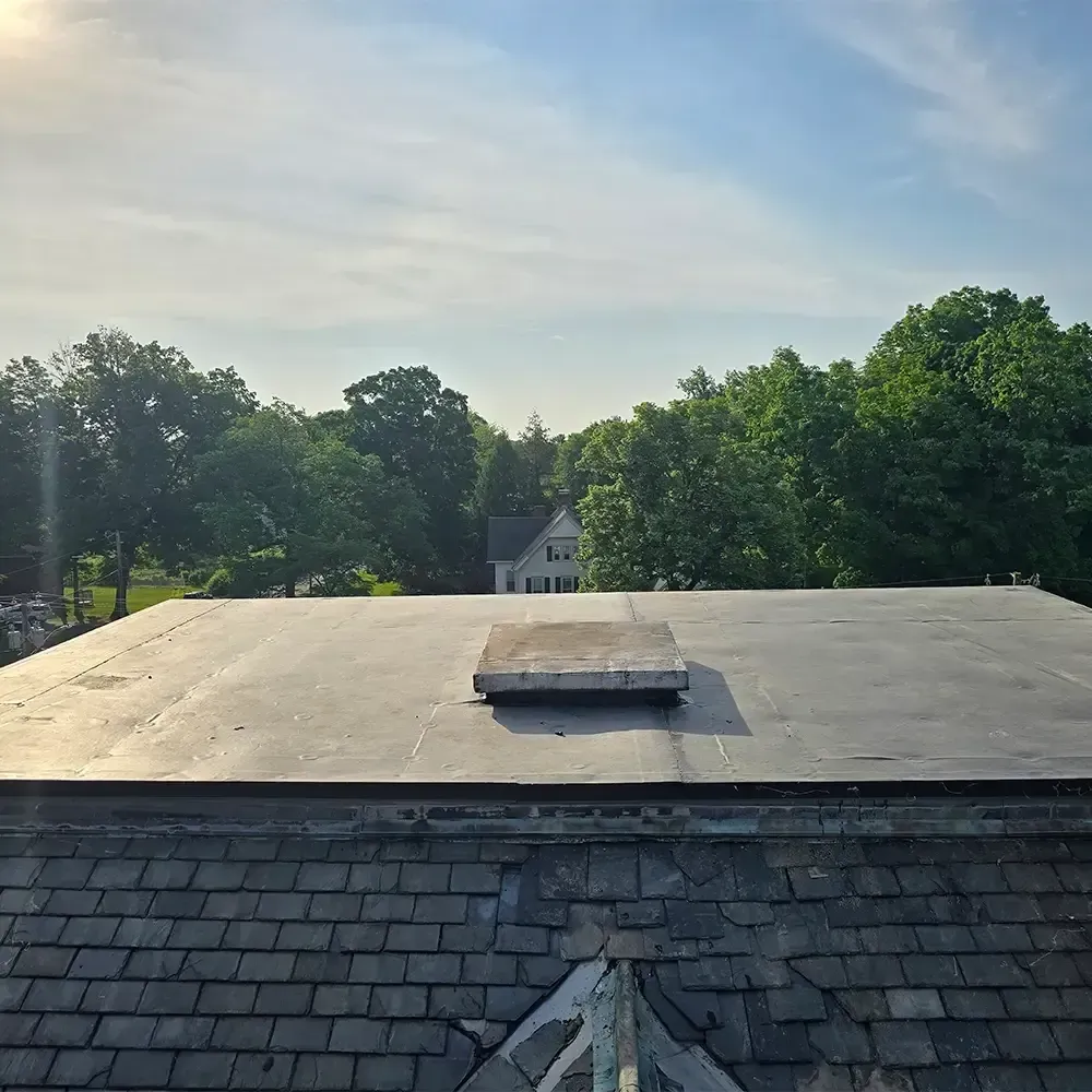 A flat rubber-membrane roof with a small skylight, viewed from above a slate-tiled roof against a sunny, wooded backdrop.