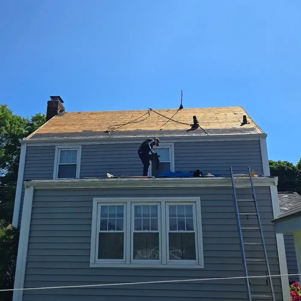 A worker on the sloped roof of a blue-sided house with a lower flat roof and a tall metal ladder leaning against the side.