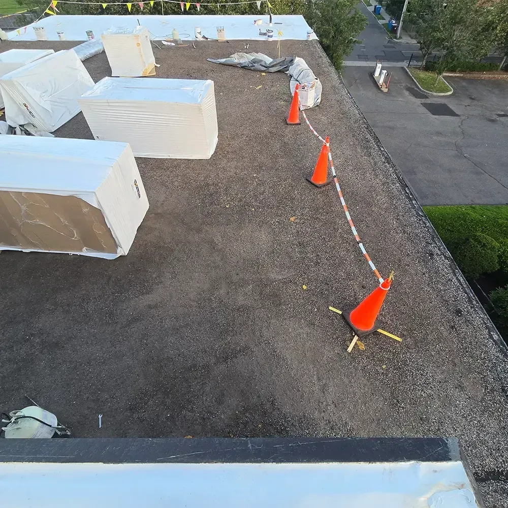 A flat commercial rooftop under construction, featuring white material pallets and orange safety cones with a caution tape.