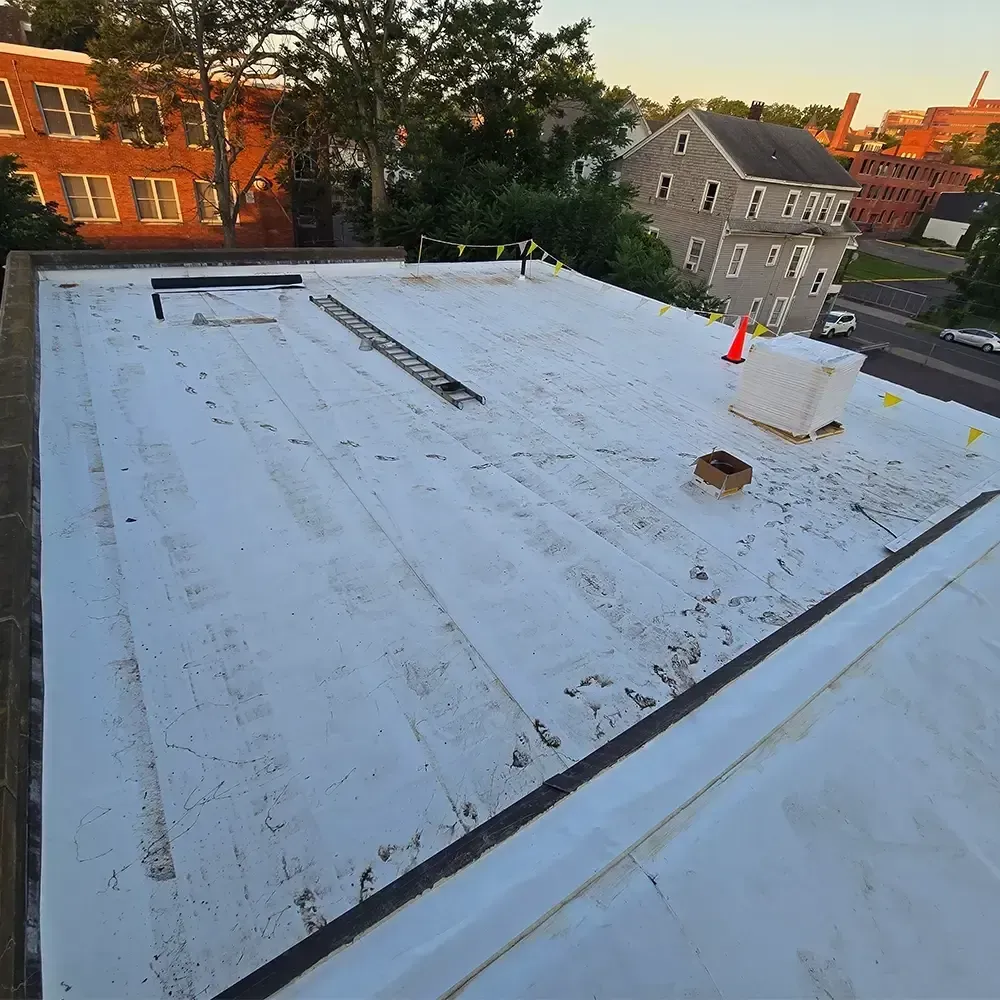 A white flat roof with a ladder, a small HVAC unit, and a caution cone, overlooking a residential neighborhood.