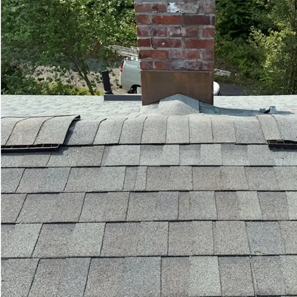 A brick chimney on a shingled roof with raised, damaged shingles near the flashing and vent.