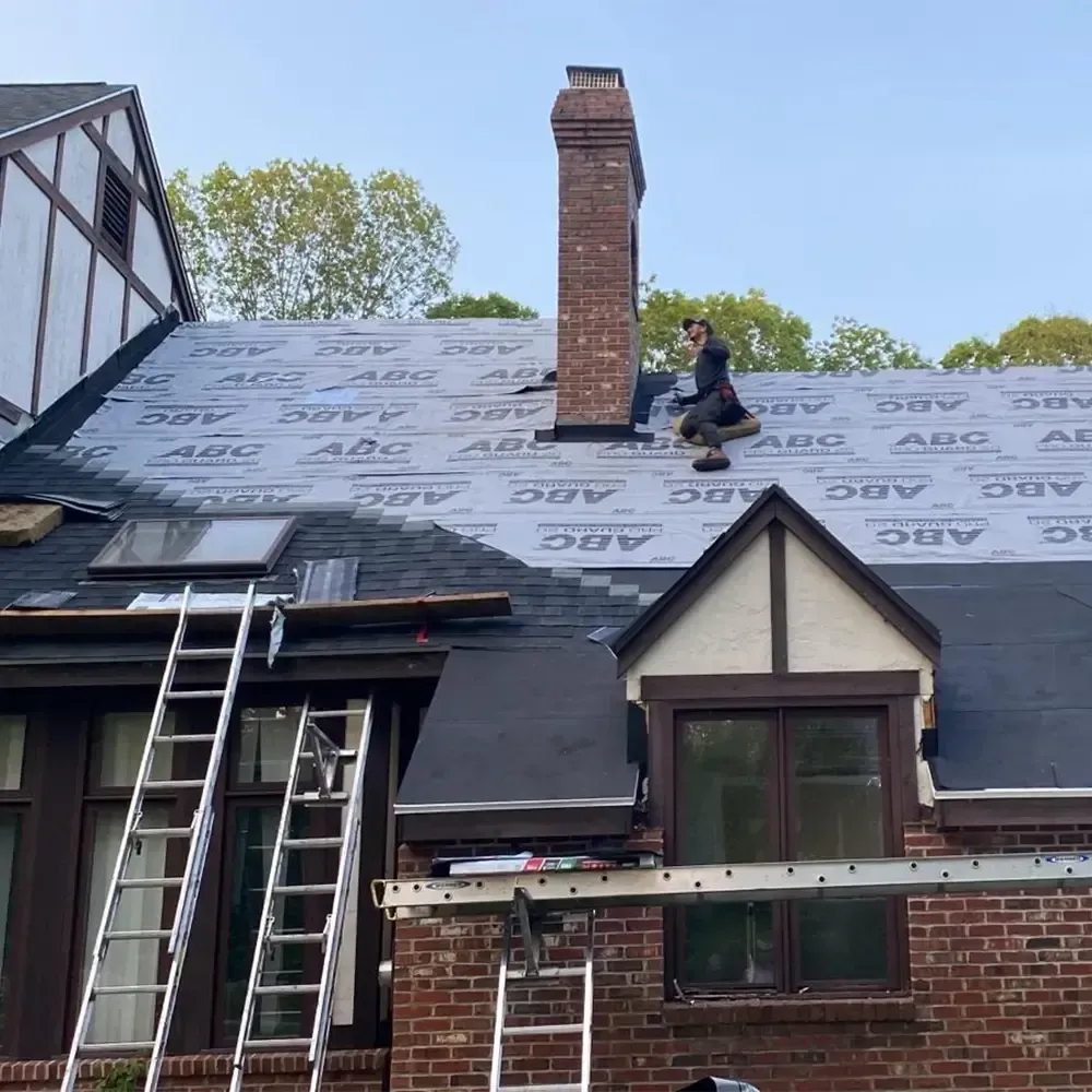 A person in dark clothing works on a roof covered in gray protective underlayment next to a brick chimney.
