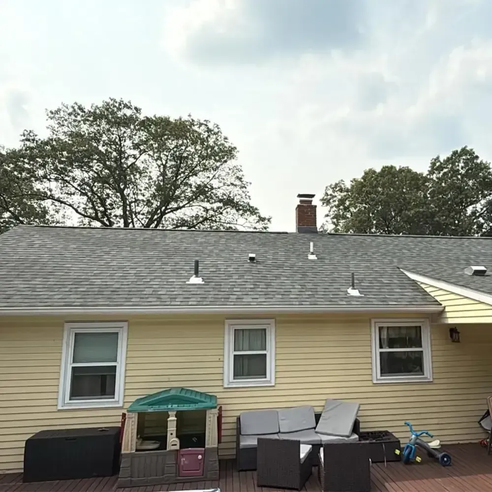 A yellow house with a gray shingle roof features a backyard deck with patio furniture and a children's playhouse.