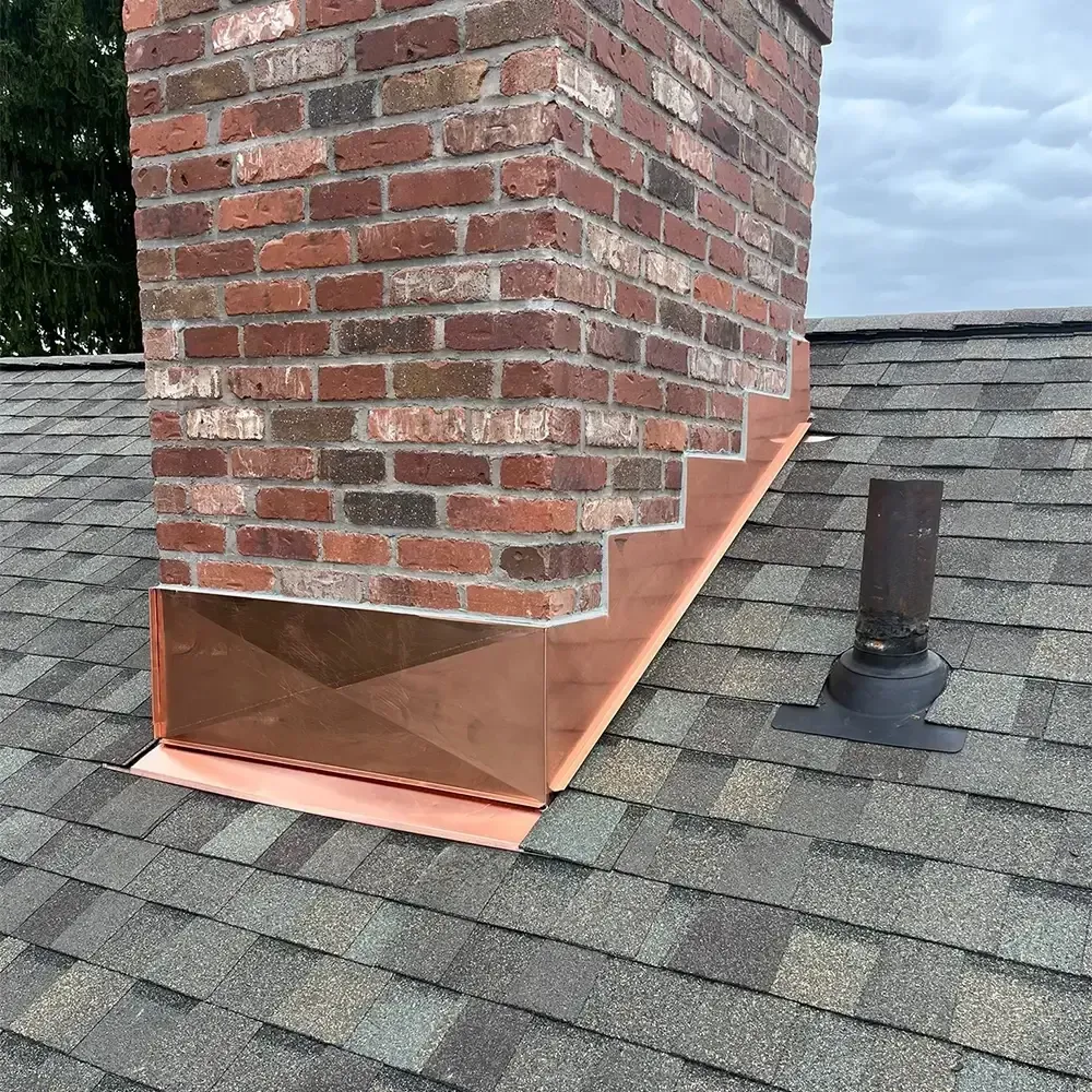 A copper chimney flashing installed on a shingled roof with a black plumbing vent pipe nearby.