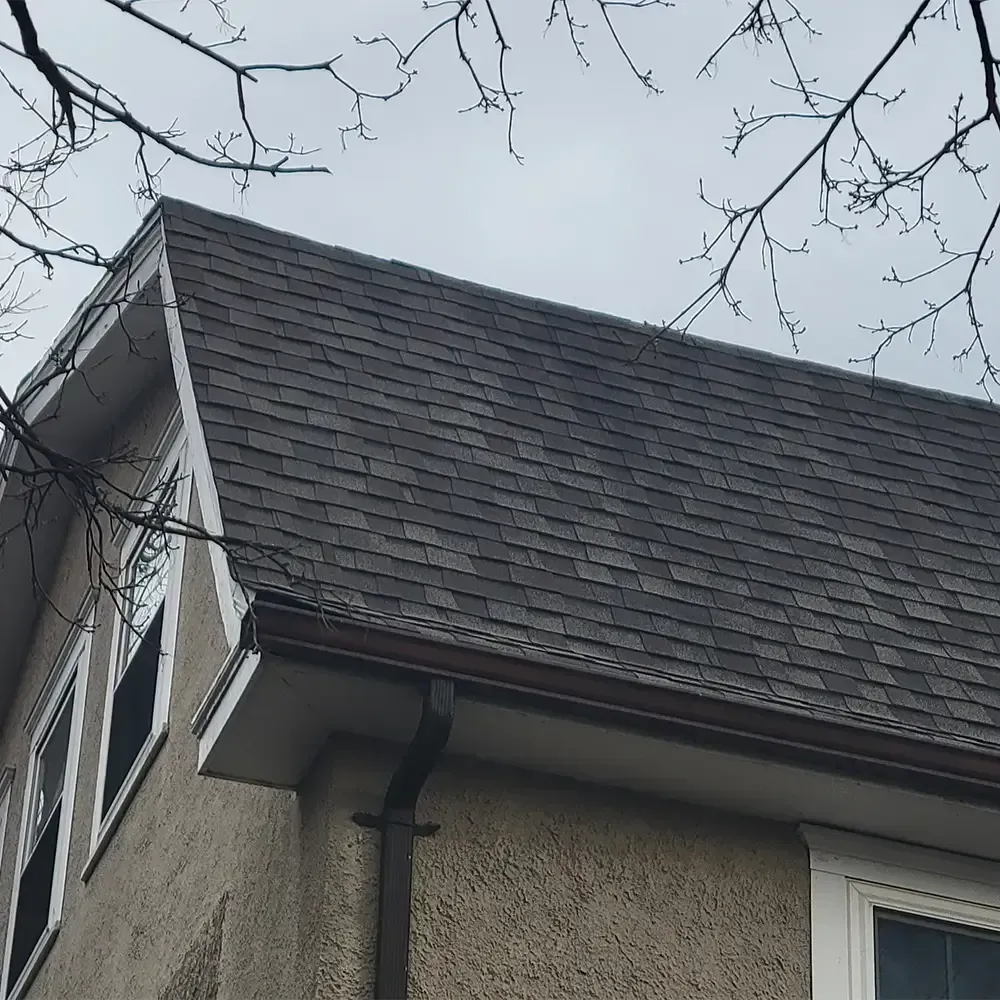 A side view of a house exterior showing a dark gray shingled roof, brown gutters, a downspout, and textured beige siding.