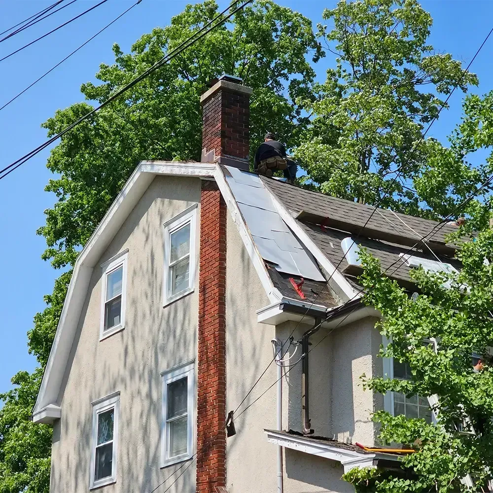 A person works on the roof of a beige house with a brick chimney and metal roof sections on a sunny day.