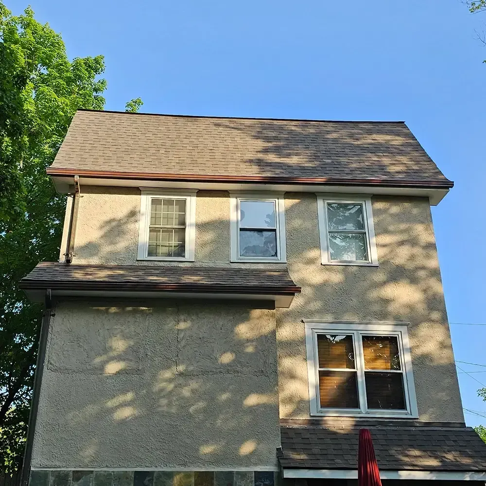 A tan stucco house with brown shingles, dark-framed windows, and trees on the left against a clear blue sky.