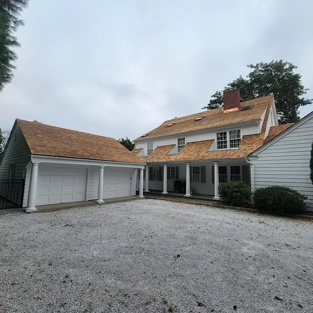 A white house with a newly installed cedar shingle roof and a matching detached two-car garage sits on a gravel driveway.