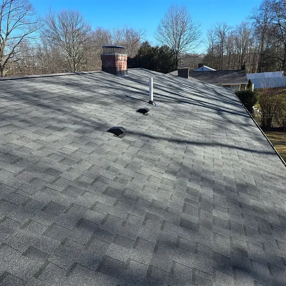 A view looking down a gray asphalt shingle roof with a brick chimney and two vents under a clear blue sky.