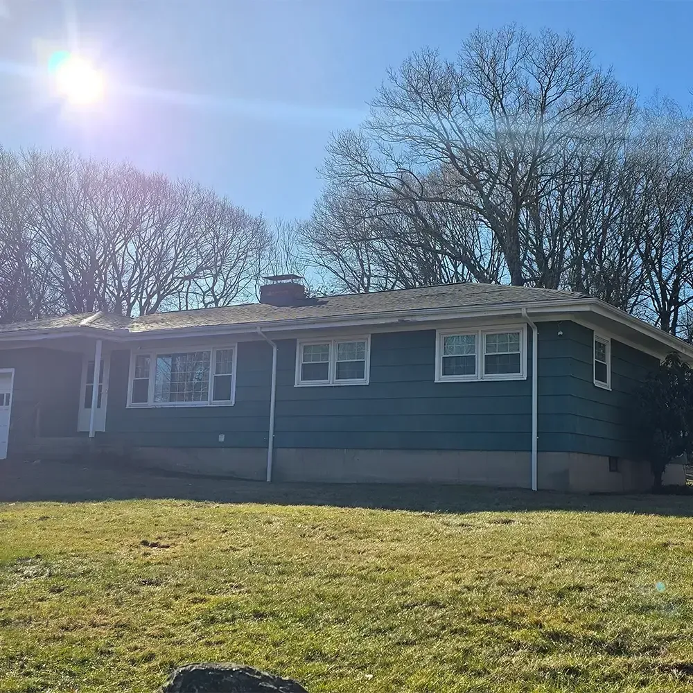 A teal, single-story house with a white trim and chimney, set against a sunny sky with bare trees in the background.