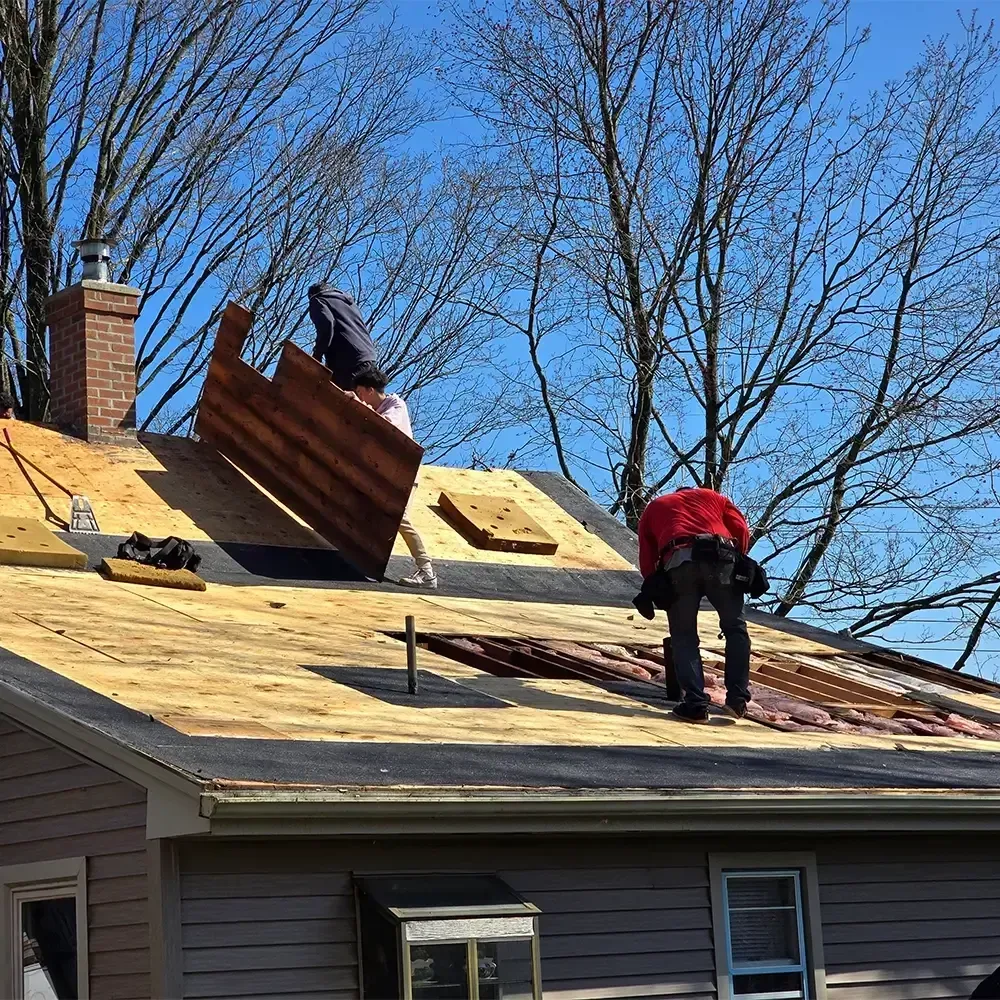 Two workers installing plywood panels on a residential roof against a bright blue sky.