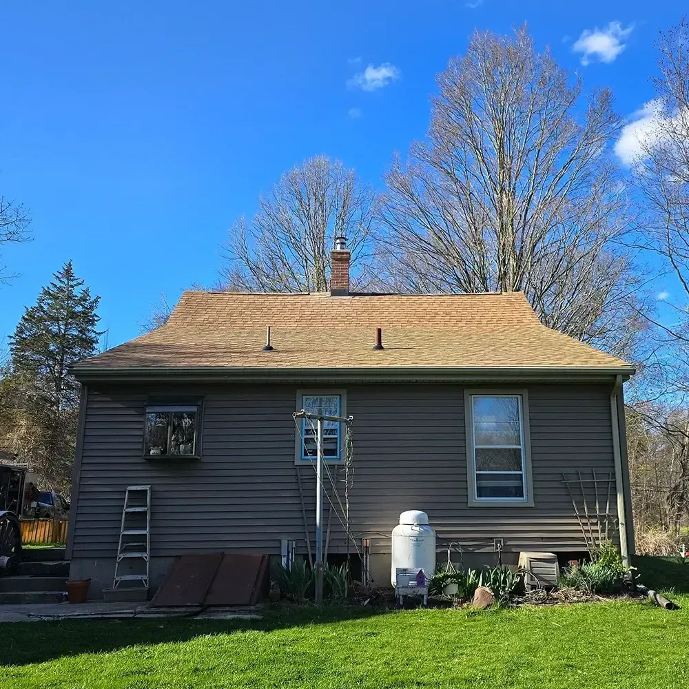 A gray, one-story house with a brown shingled roof, two windows, a ladder, and a propane tank in a grassy yard.