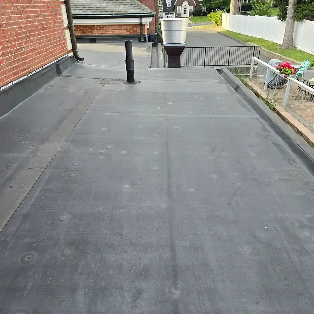 A flat, black-rubberized rooftop with a vent pipe, a brick wall on the left, and a white fence visible in the background.