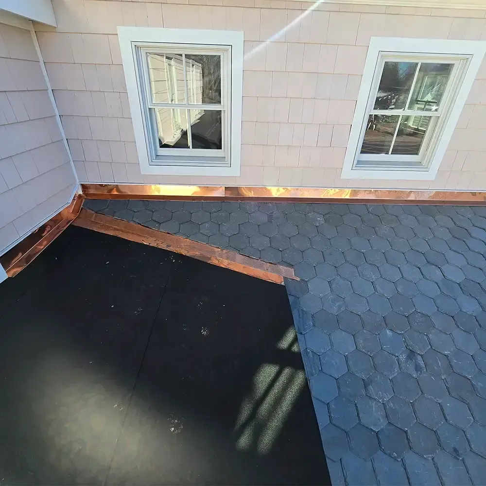 A low-angle view of a roof with black hexagonal shingles and a flat, dark membrane section, bordered by copper flashing.