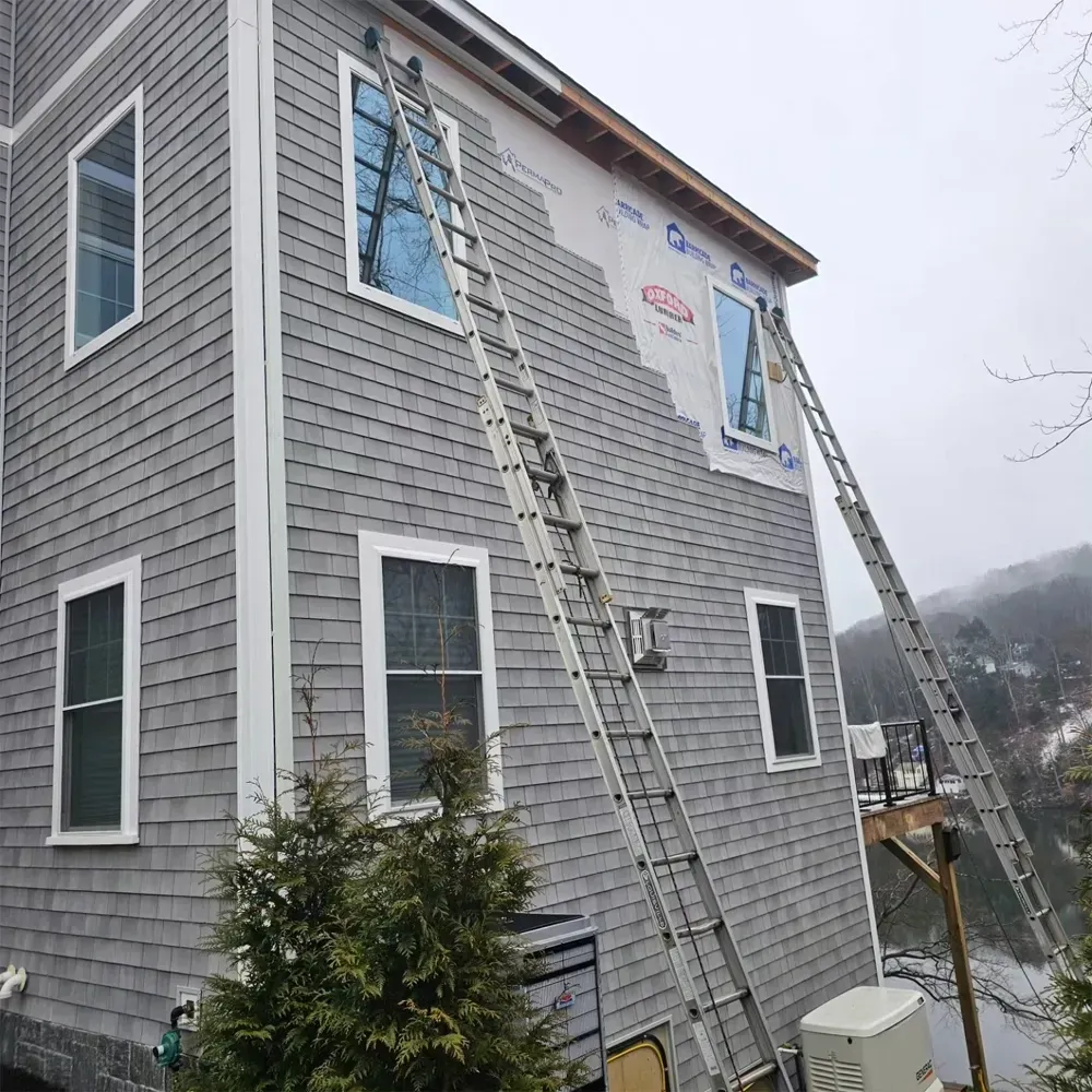 Two ladders lean against the gray-shingled wall of a house under construction, with exposed building wrap near the roof.