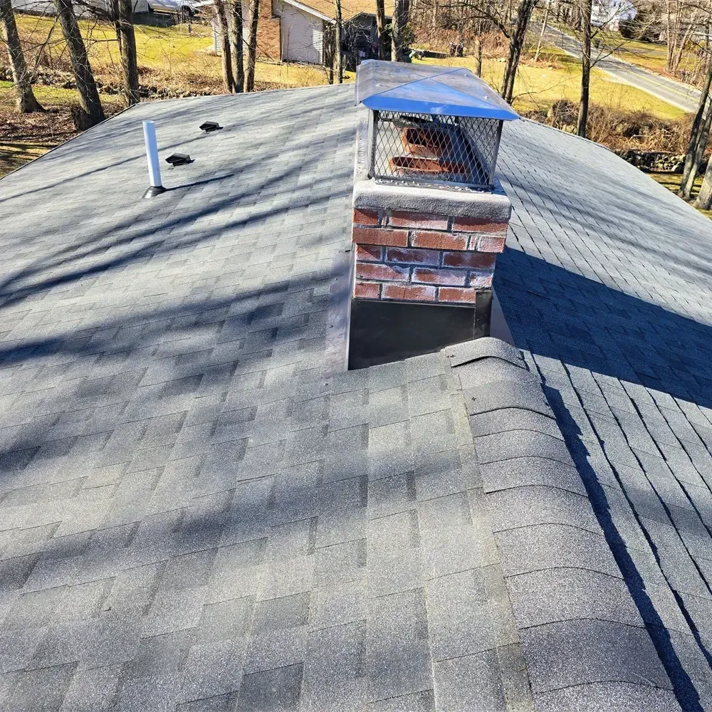 A gray shingled roof features a brick chimney with a metal cap and a white vent pipe in a sunny outdoor setting.